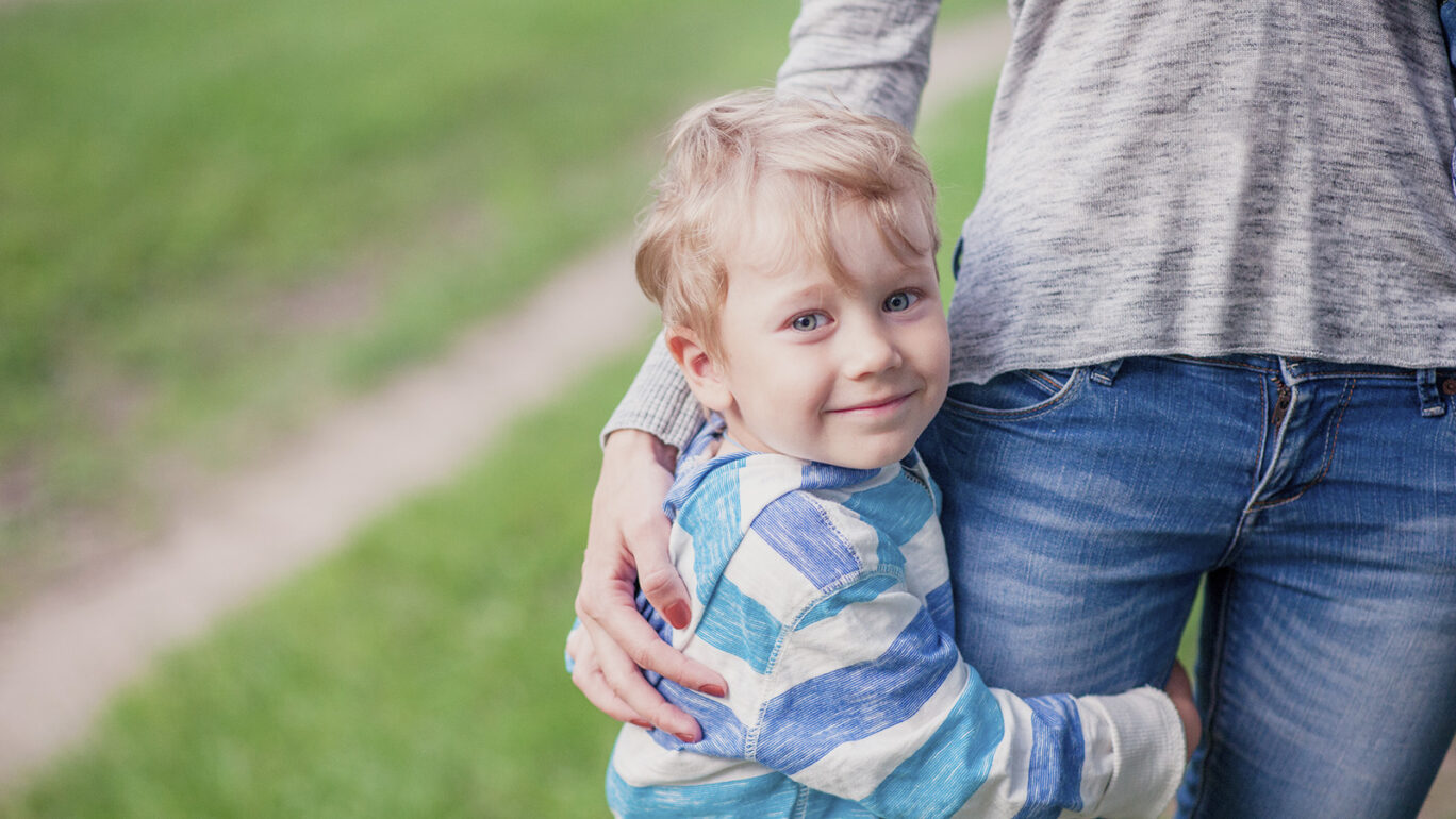 Poika katsoo hymyillen kameraa ja halaa naisen jalkaa