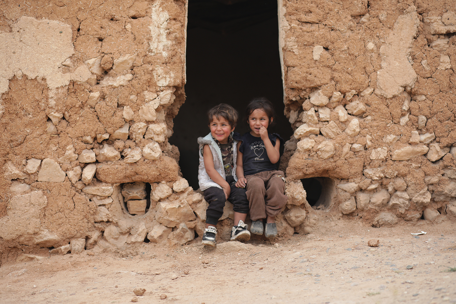 Kaksi lasta istuu hymyillen kivestä ja hiekasta tehdyn talon ovella.