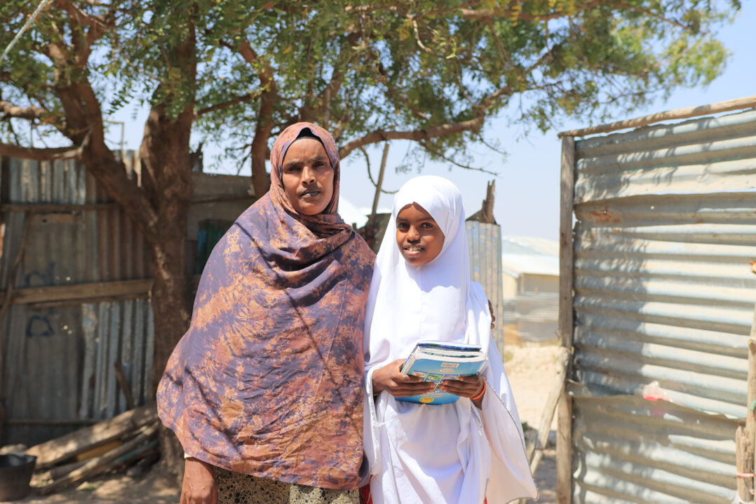 Aikuinen nainen ja teini-ikäinen tyttö seisovat ulkona peltiaidan edessä. Tytöllä on koulukirjoja kädessä.