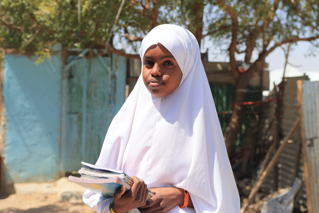 Teini-ikäinen tyttö seisoo ulkona peltisen aidan edessä käsissään koulukirjoja.