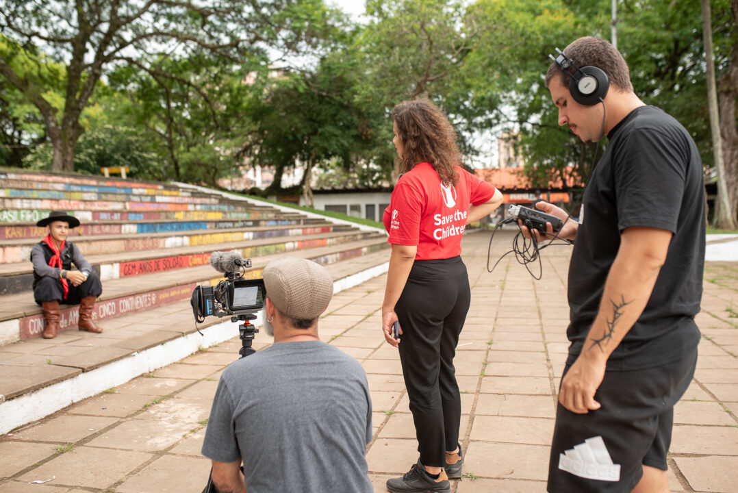 Nuori istuu portailla ja häntä on kuvaamassa kaksi mieshenkilöä. Kuvassa myös nainen, jolla on Save the Children -tekstillä varustettu paita päällä. 