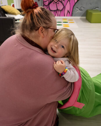 Äiti ja lapsi halaavat toisiaan sisäleikkipaikalla. Lapsi katsoo hymyillen kameraan.