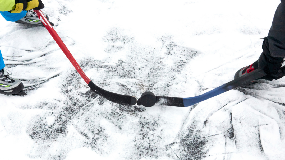 Kaksi henkilöä pelaa jääkiekkoa ulkona lumisella jäällä. Kuvassa näkyy kaksi jääkiekkomailaa, punainen ja sininen, jotka koskettavat kiekkoa keskellä. Pelaajien luistimet ja osa jaloista näkyvät, ja jäässä on paljon luistimen jälkiä.