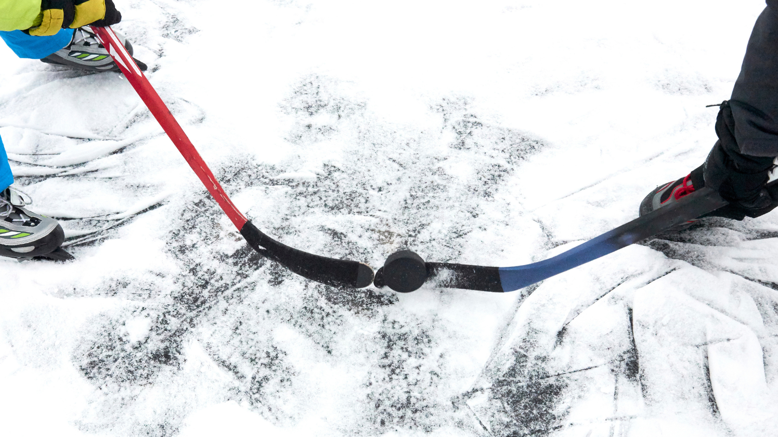 Kaksi henkilöä pelaa jääkiekkoa ulkona lumisella jäällä. Kuvassa näkyy kaksi jääkiekkomailaa, punainen ja sininen, jotka koskettavat kiekkoa keskellä. Pelaajien luistimet ja osa jaloista näkyvät, ja jäässä on paljon luistimen jälkiä.