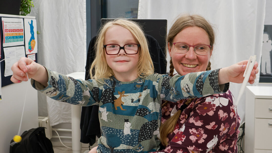 Poika istuu äitinsä sylissä ja katsoo suoraan kameraan. Hänellä on kädet sivuille levitettynä ja käsissään kynä ja lelu. Äiti hymyilee taustalla.