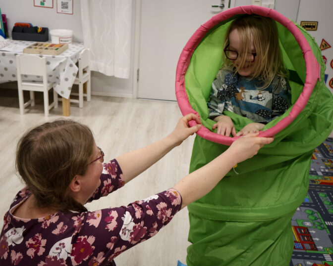 Äiti pitää kiinni leikkitunnelista ja poika kurkistaa hymyillen tunnelista ulos kohti äitiä.