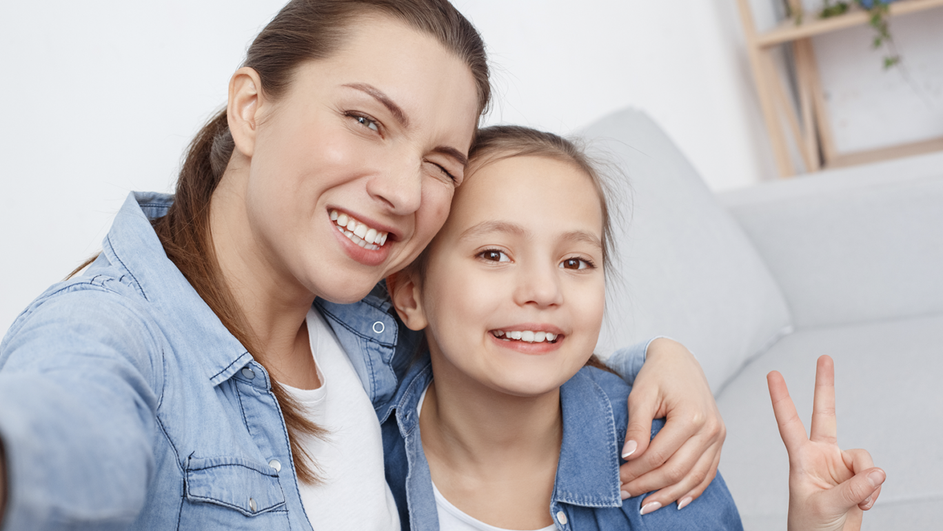 Nainen ja tyttö istuvat sohvalla ja virnistävät hymyillen