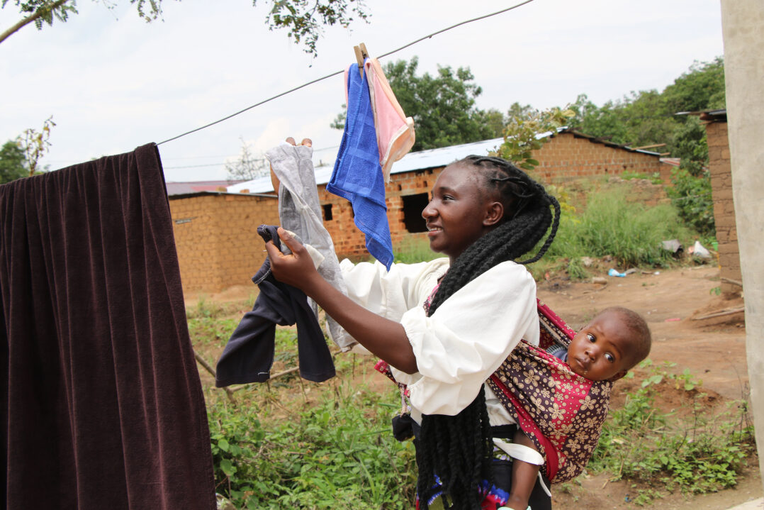 Nainen vauva kantorepusta kurkistaen ripustaa pyykkejä narulle ulkona.