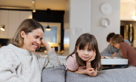Nainen istuu sohvalla ja katsoo hymyillen vieressä olevaa lasta. Taustalla poika ja mies lukevat pöydän ääressä.
