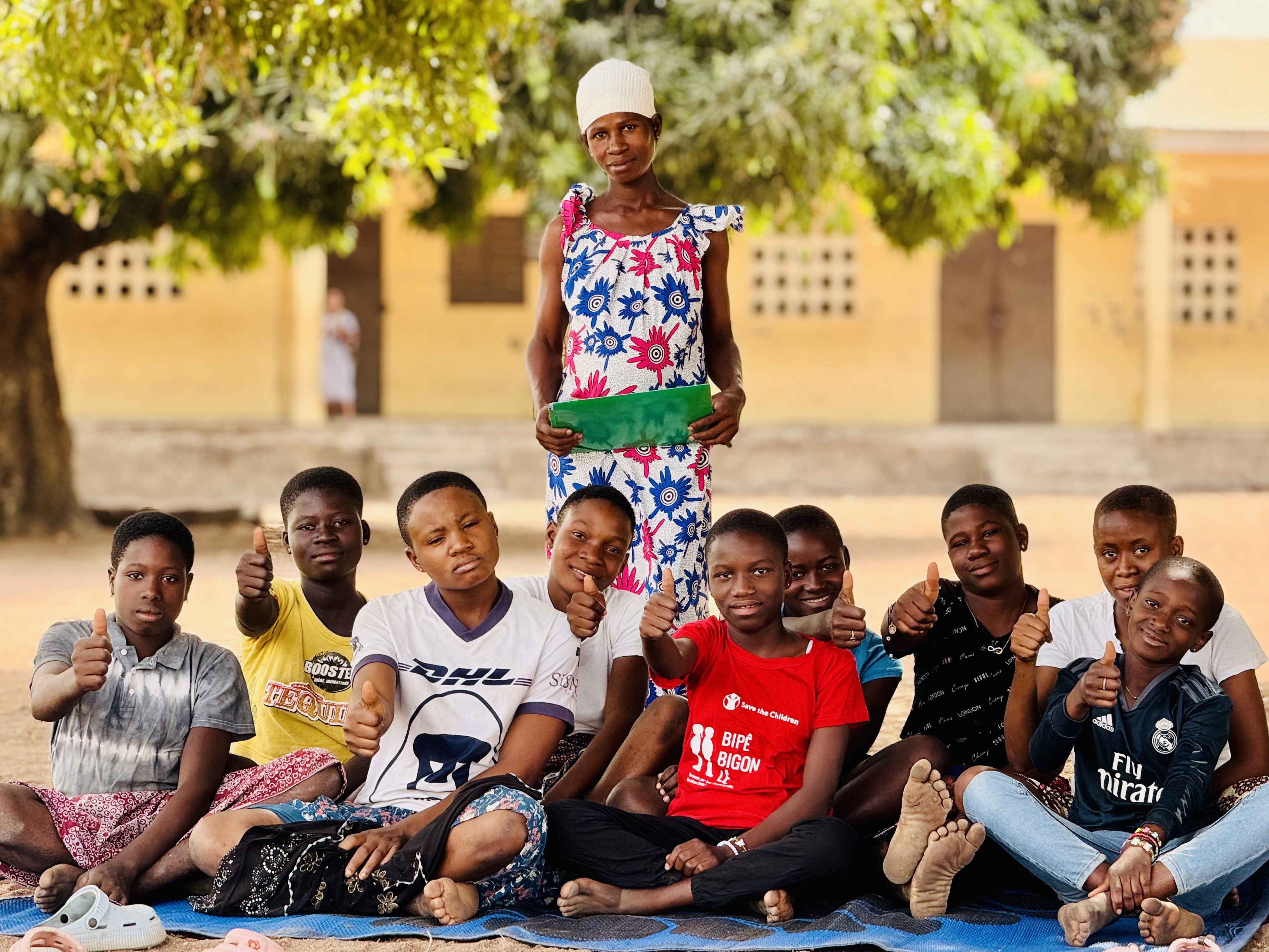 Nuoria istuu hiekkaisella maalla viltin päällä. Taustalla seisoo aikuinen.