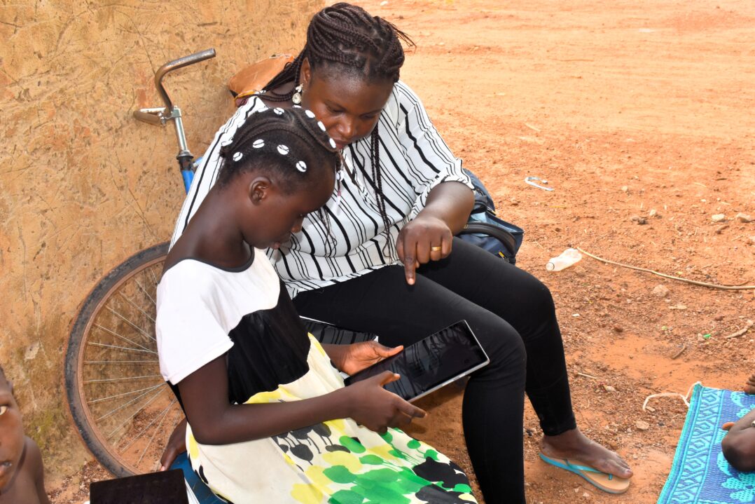 Tyttö katselee naisen kanssa tablettia. Taustalla hiekkainen maa ja polkupyörä. 