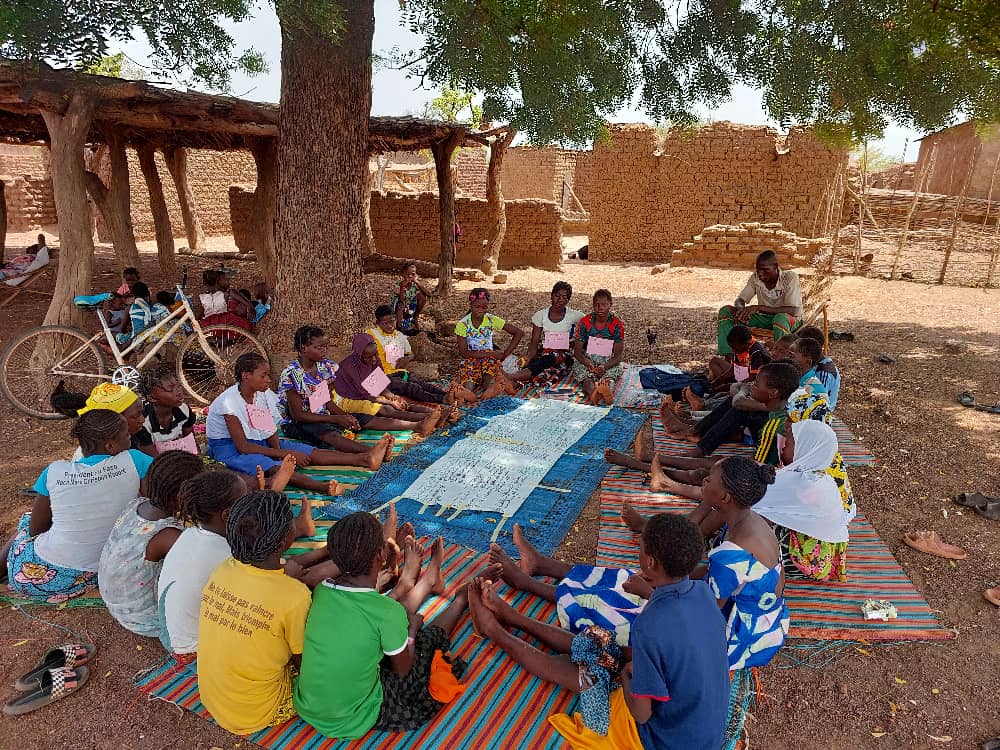 Ihmisiä istumassa puun alla varjossa vilttien päällä ringissä. Taustalla hiekkaista maata. 