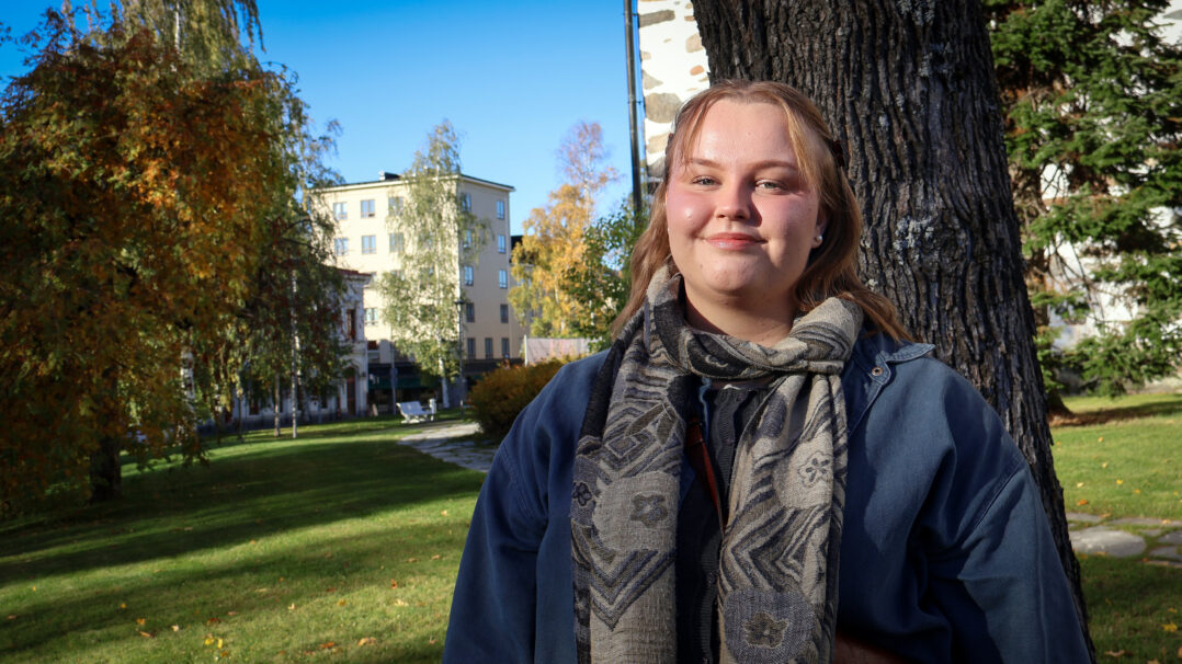 Tukihenkilö Helmi Romppanen seisoo puistomaisessa ympäristössä suuren puun vieressä. Hänellä on yllään sininen takki ja huivi, ja taustalla näkyy syksyinen nurmialue, puita ja kerrostaloja.