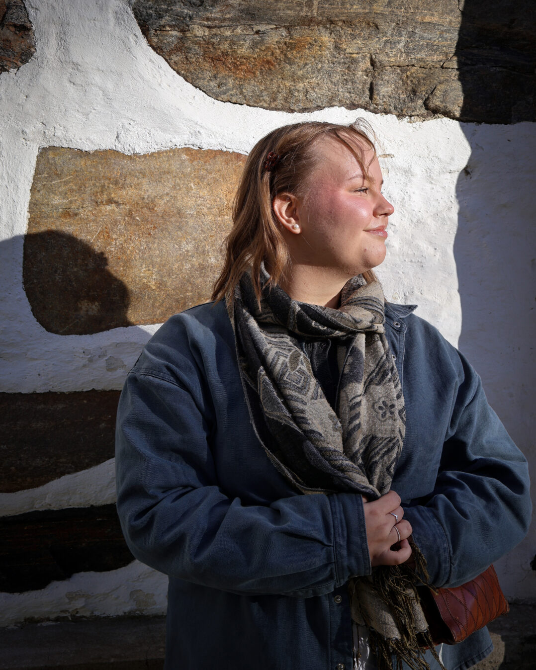 Tukihenkilö Helmi Romppanen seisoo ulkona hirsiseinän edessä, yllään sininen takki ja huivi. Hän katsoo sivulle ja nojaa rauhallisesti seinään luonnonvalossa.