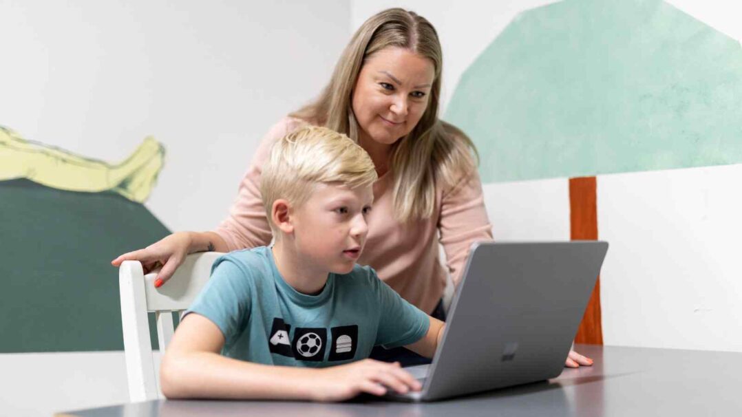 Lapsi tietokoneella pöydän ääressä, vanhempi seisoo taustalla nojautuneena katsomaan ruutua.