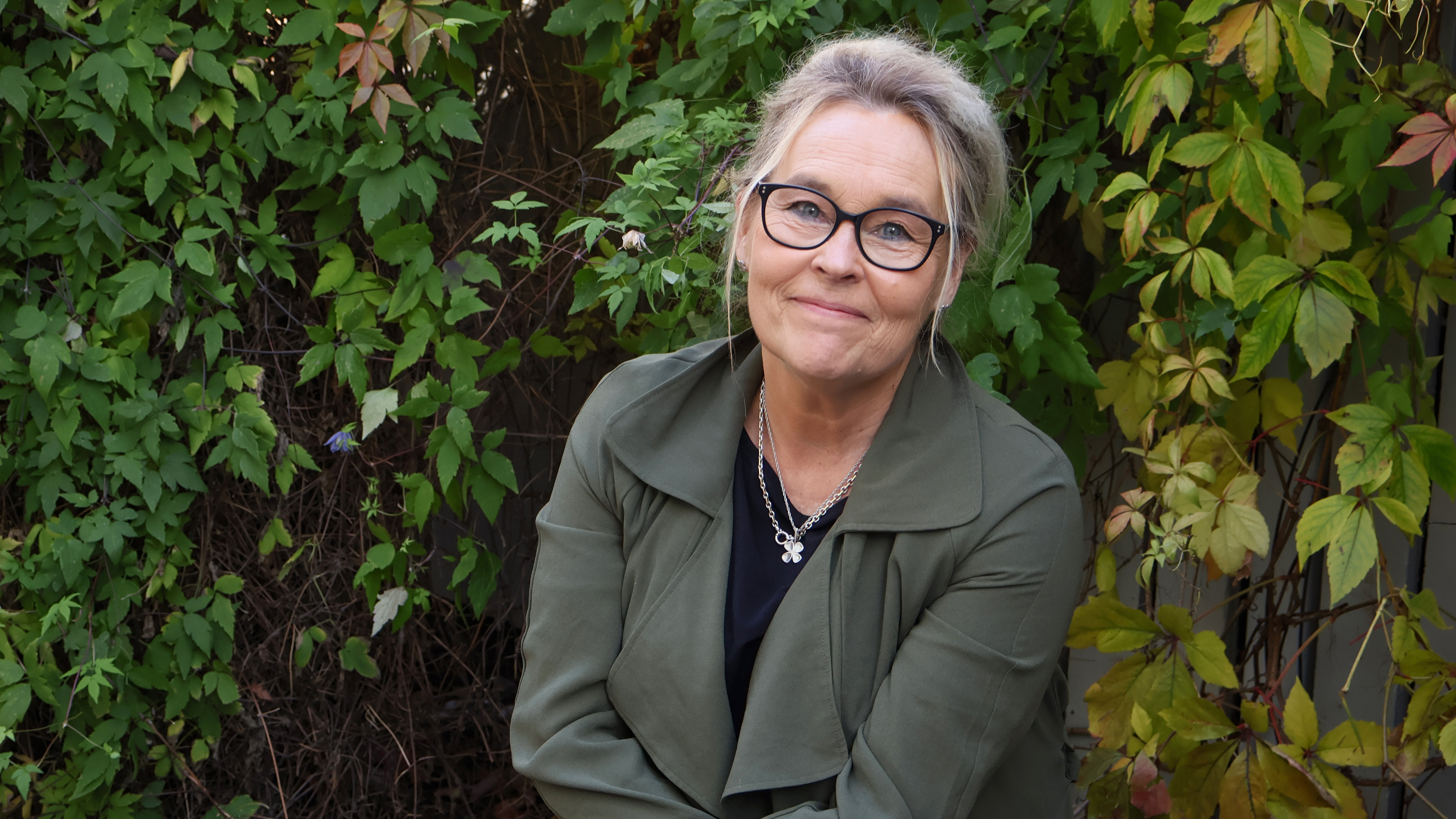 Nainen katsoo kameraan hymyillen. Taustalla vihreän kasvillisyyden peittämä seinä.