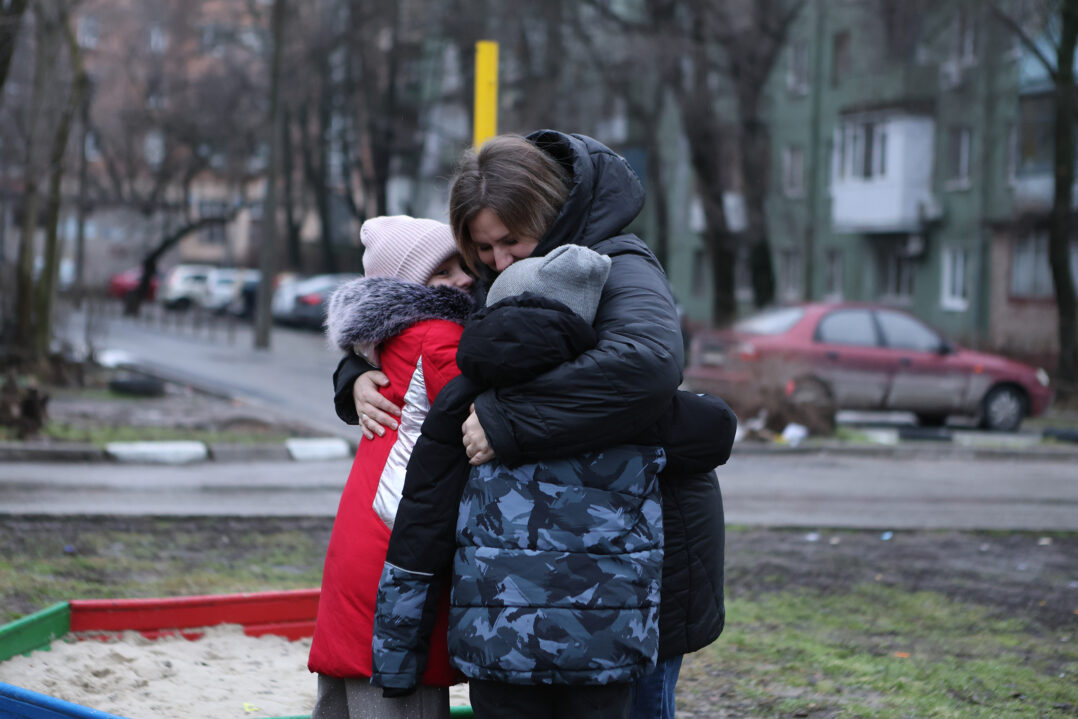 Äiti halaa kahta lastaan tiukasti. He seisovat ulkona talvivaatteet päällä.