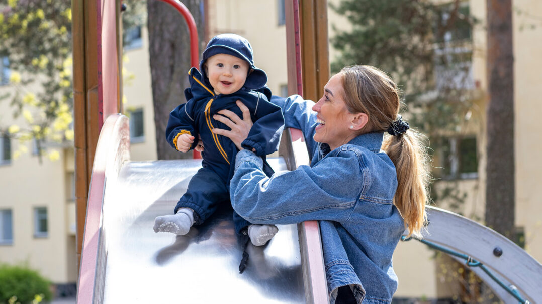 Äiti auttaa hymyillen pientä lasta laskemaan liukumäkeä