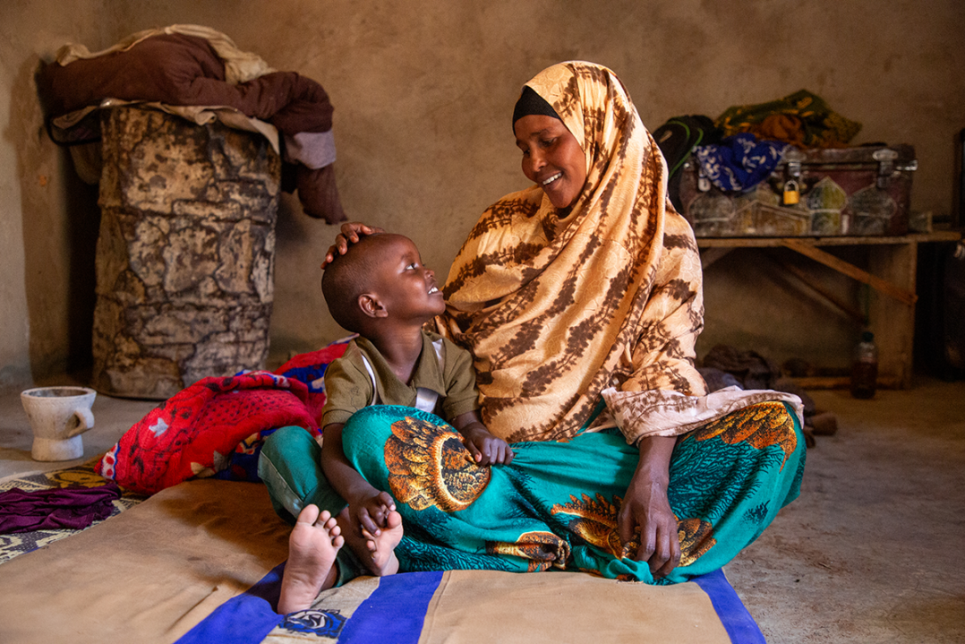 Äiti ja lapsi istuu vierekkäin lattialla. Taustalla kankaita ja tavaroita. 