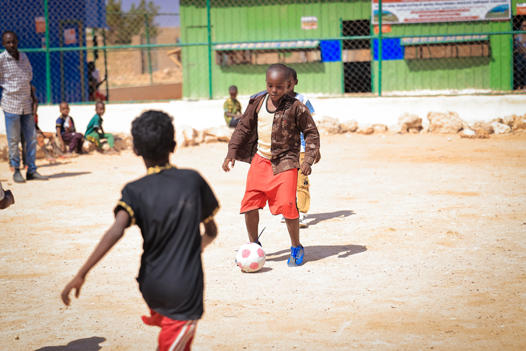 Poika pelaa jalkapalloa hiekkakentällä. Edessä toinen lapsi.