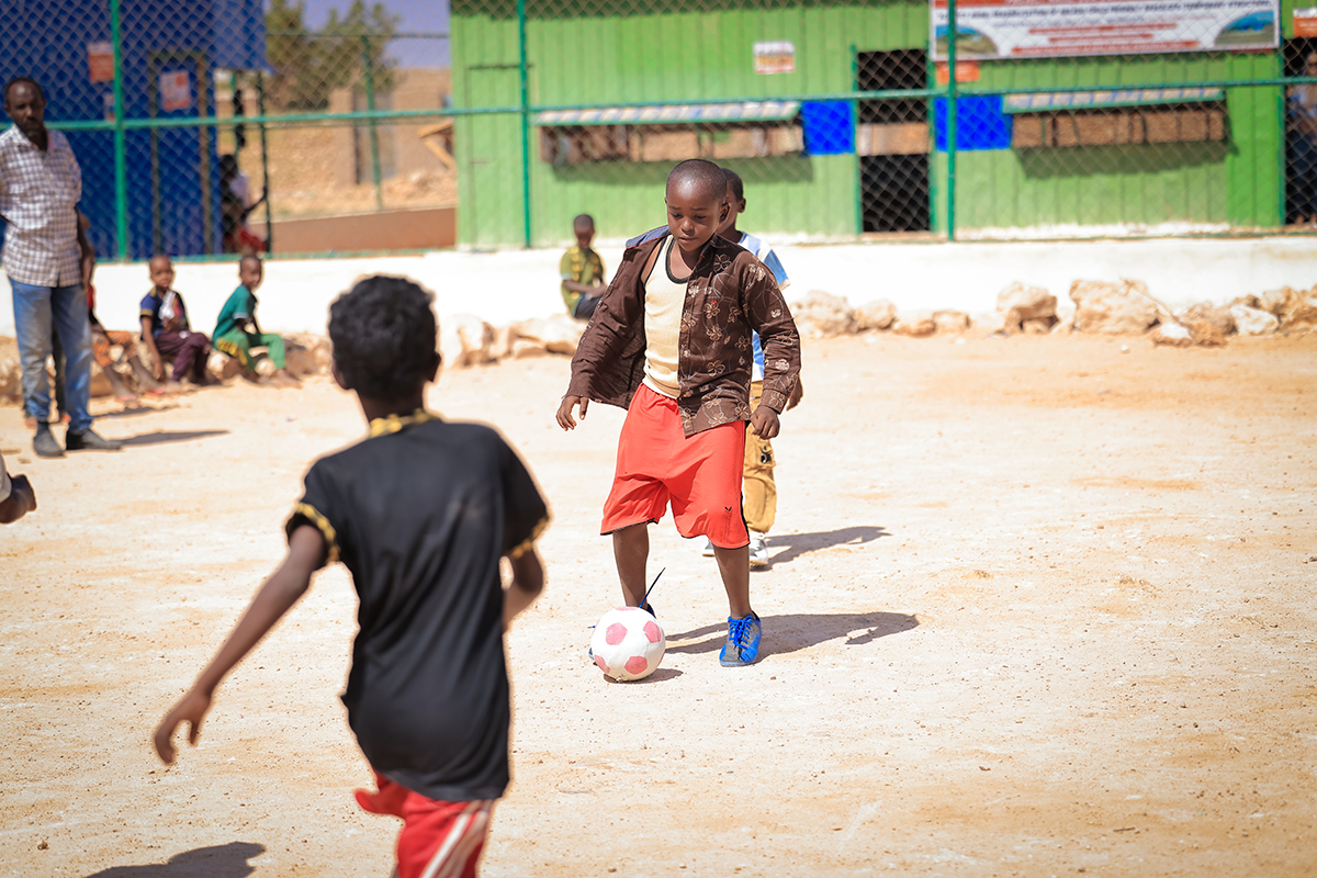 Poika pelaa jalkapalloa hiekkakentällä. Edessä toinen lapsi.