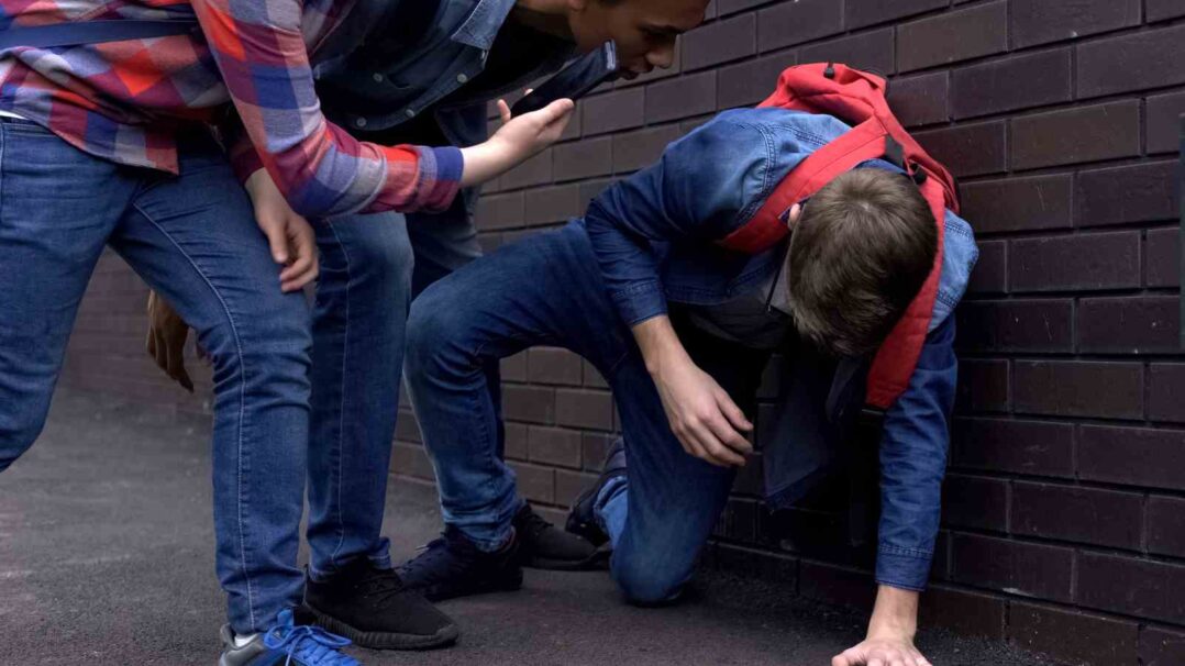 Lapsi ottaa tukea maasta, kiusaajat ovat kumartuneena hänen puoleensa ja kuvaavat puhelimella.