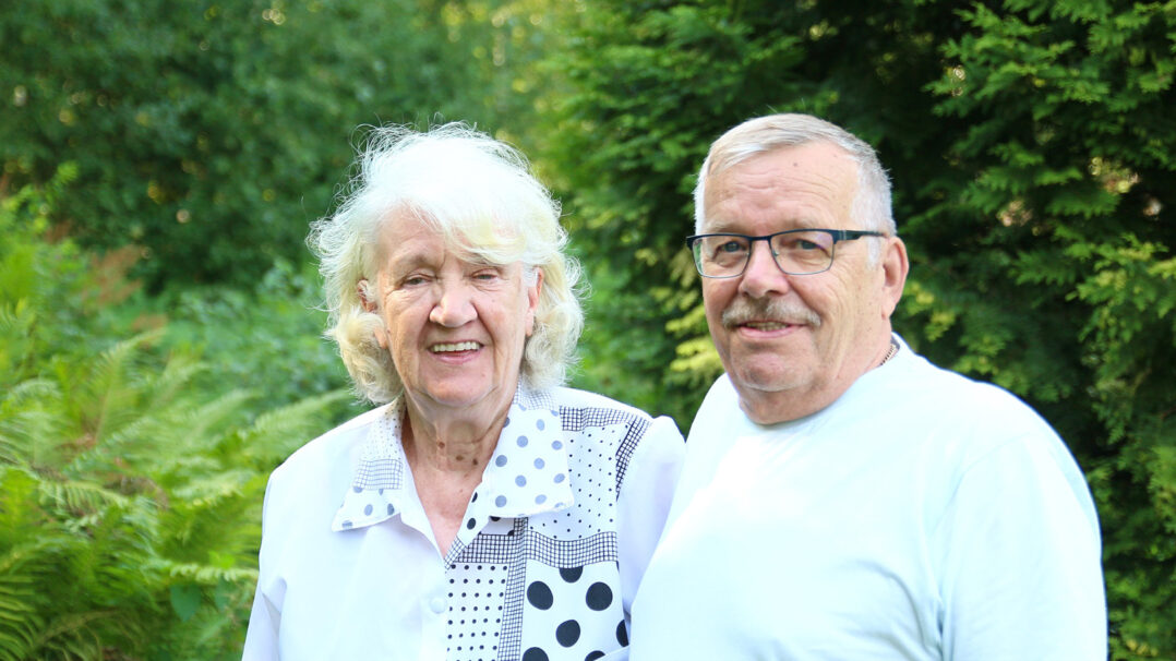 Nainen ja mies seivovat vierekkäin ja hymyilevät. Taustalla vihreää pensasta ja puita.