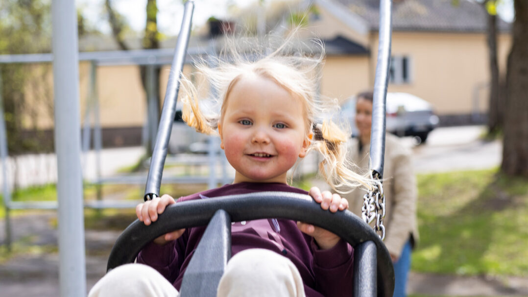 Lapsi keinuu ja hymyilee kuvassa. Taustalla nainen