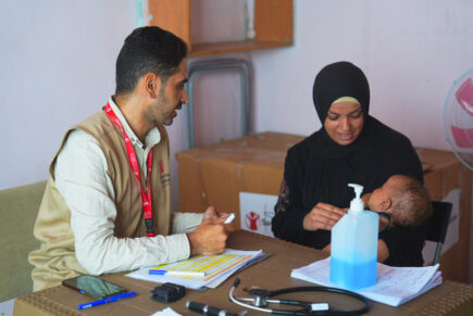 Nainen istuu vauva sylissään pöydän ääressä. Mies istuu toisella puolella pyötää ja kirjoittaa paperille.