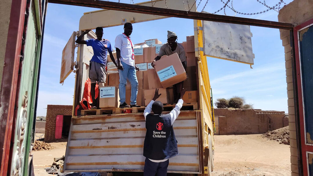 Miehiä purkamassa laatikoita kuorma-autosta. Taustalla hiekkaista maata. Miehillä Save the Children -liivi päällä.