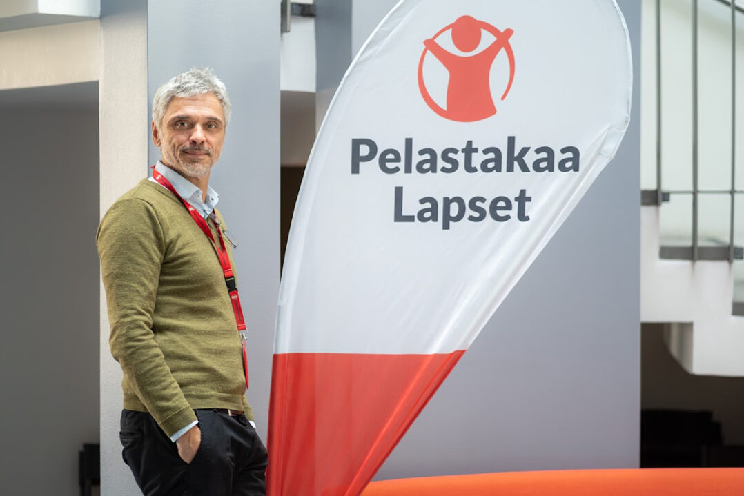 Mies seisoo nojaten seinään. Vieressä Pelastakaa Lapset -banderolli.