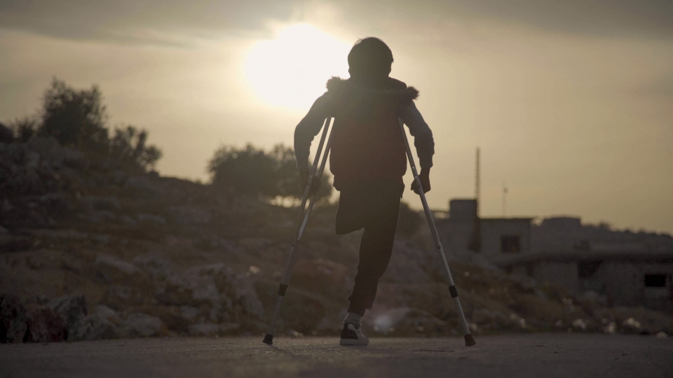 Lapsi seisoo kainalosauvojen kanssa vasten aurinkoa, toinen jalka on amputoitu.