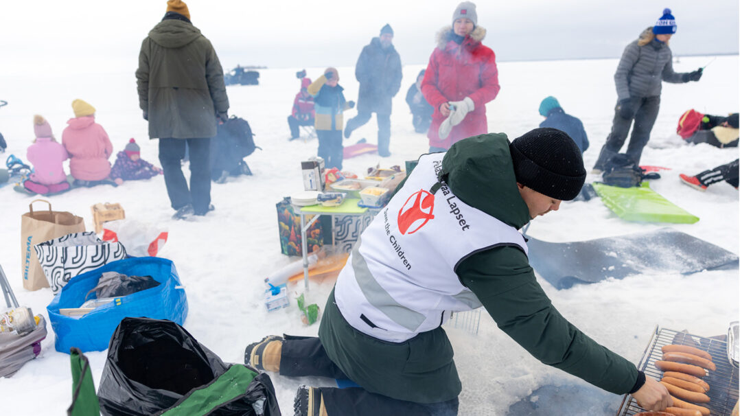 Pelastakaa Lasten työntekijä paitsaa makkaroita. Taustalla joukko vapaaehtoisia ja lapsia jäällä.