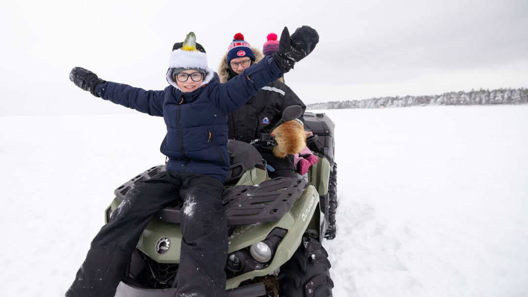 Poika ja aikuinen istuvat mönkijän päällä jäällä. Poika levittää kätensä sivuille ja hymyilee