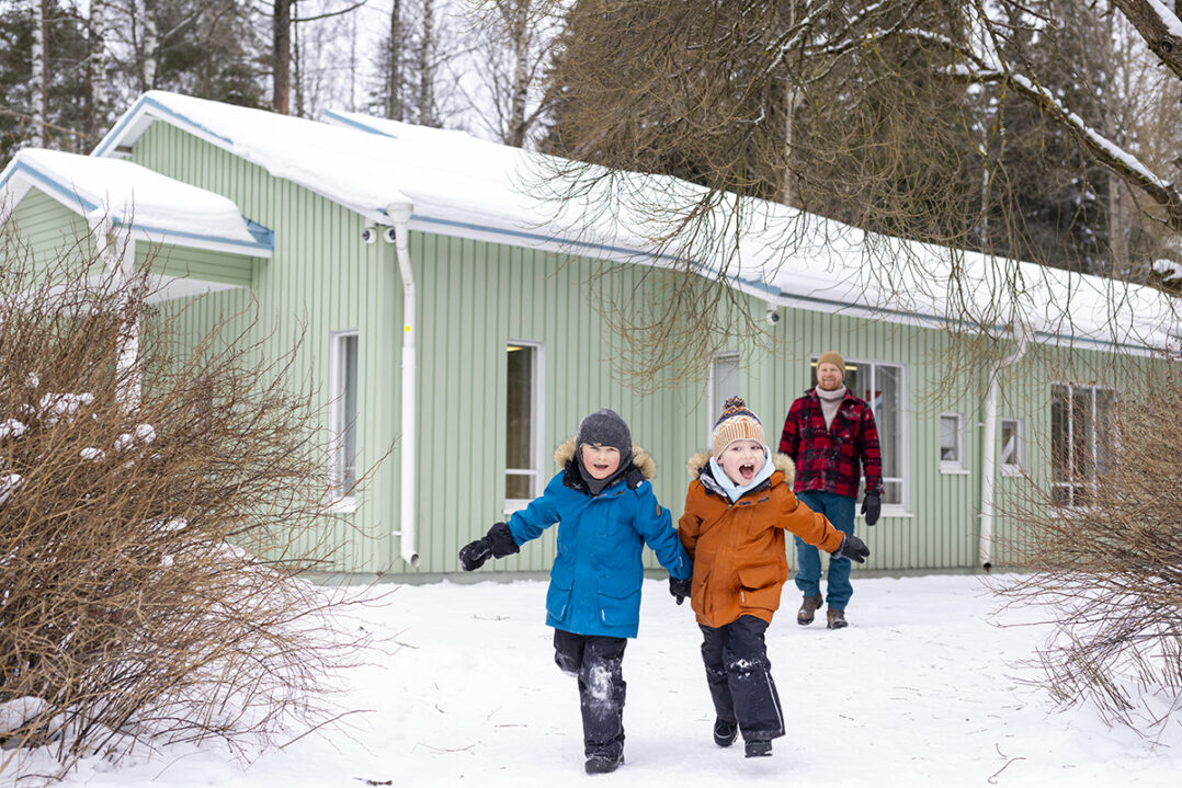 Talvimaisema, jossa kaksi talvivaatteisiin pukeutunutta lasta juoksee lumessa kohti kameraa. Taustalla aikuinen mieshenkilö ja puuverhoiltu vihreä talo