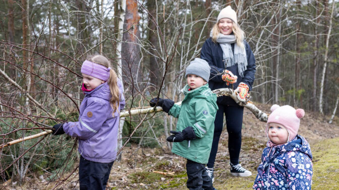 Aikuinen ja kolme lasta kantavat kaatunutta puunrunkoa metsässä